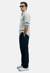 Helle beigefarbene kurzärmelige Pullover, marinefarbene Hose, weiße Sneaker, über einem blau-gestreiften Hemd getragen, mit Sonnenbrille.