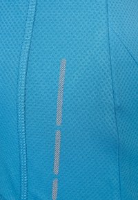 Světlě modrý sportovní materiál s texturovaným vzorem, s reflexními stříbrnými pruhy a viditelnými švy pro větší detail.