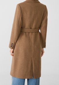 Manteau en laine brun avec ceinture porté sur un jean bleu, vu de dos, doté de poignets à boutons et d'une fente à l'arrière.