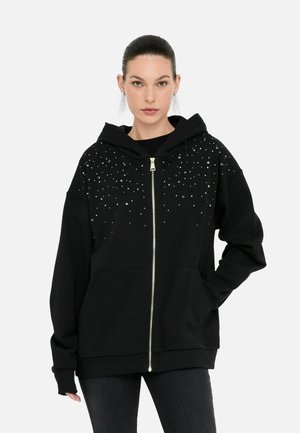 Modella che indossa una felpa nera con zip decorata con brillanti, in piedi davanti a uno sfondo bianco puro.