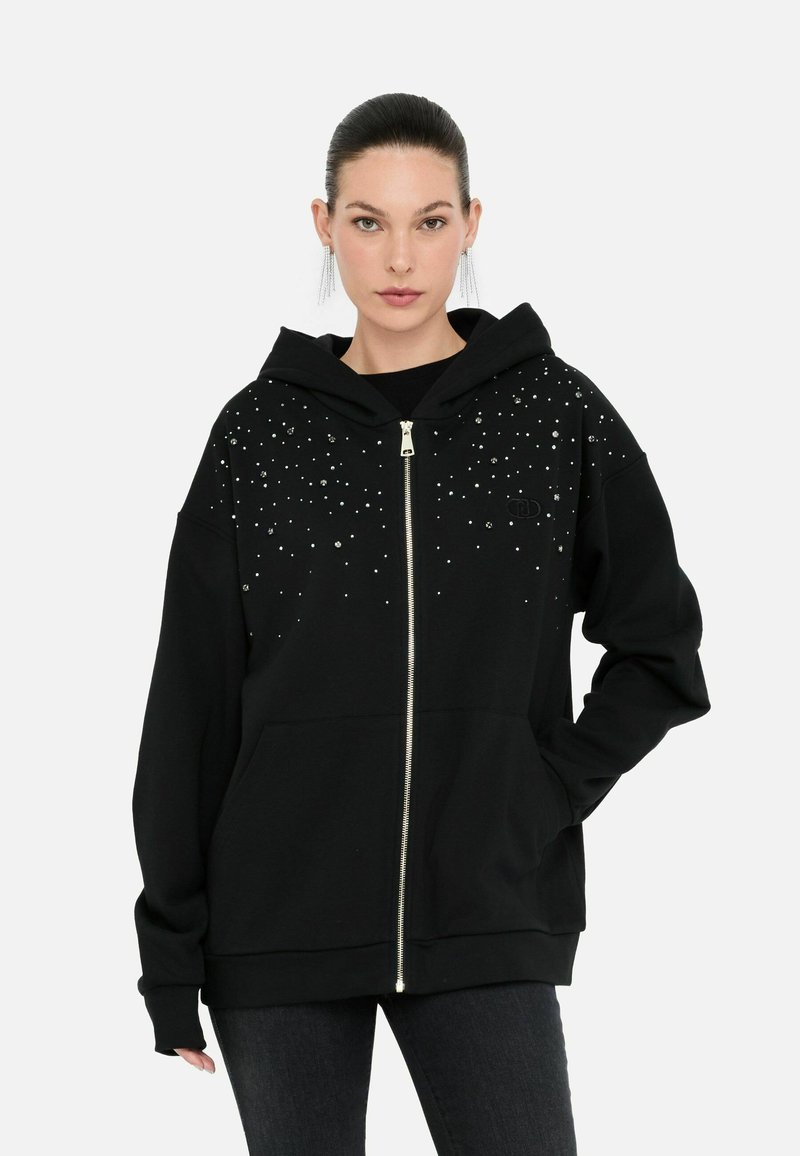 Modèle portant un hoodie zippé noir orné de décors scintillants, se tenant devant un fond blanc uni.
