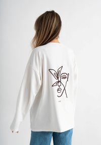 Crème lange mouw sweatshirt met een bruine lijntekening van een gezicht en een bloemelement op de achterkant, gemaakt van zachte stof.