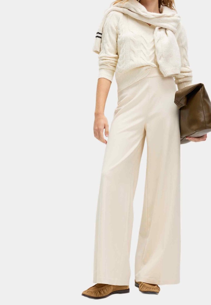 Femme portant un pantalon large crème, un pull en maille torsadée crème avec un pull assorti noué sur les épaules, des ballerines beige, tenant un grand sac marron.