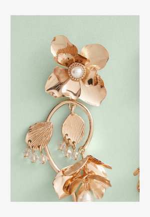 Aukso spalvos floriniai auskarai turi centrinį žiedą su perla, sudėtingus lapų akcentus ir kabinamus kristalinius karoliukus.