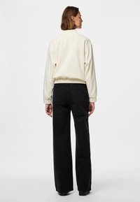 Žena smeđe kose koja nosi kremasti top dugih rukava i crne traperice visokog struka širokih nogavica stoji ispred bijele pozadine.