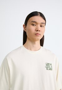 Cream-colored oversized T-shirt made of soft cotton, featuring a green illustrated logo on the left chest area and a rounded neck.