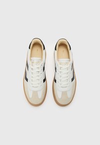 Weiße und beige Sneakers mit schwarzen Akzenten, strukturiertem Wildleder und Leder, runder Schuhspitze und flacher Gummisohle. Goldenes Logo auf der Zunge.