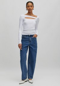 Weißes Langarmoberteil mit einem diagonalen Ausschnitt am Ausschnitt, kombiniert mit lockeren blauen Jeans und weißen spitzen Schuhen.