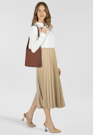 Frau in weißer Bluse und beigem, plissiertem Rock, trägt eine braune Schultertasche und beigen High-Heels, steht vor einfachem Hintergrund.