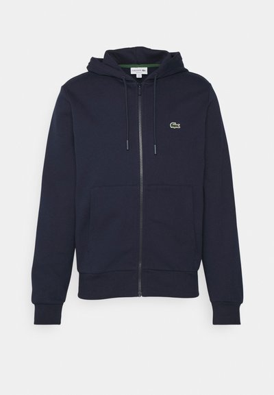 Sweat à capuche zippé bleu marine en coton doux. Il dispose d'une poche kangourou, d'une capuche à cordon de serrage et d'un petit logo vert Lacoste sur la poitrine.