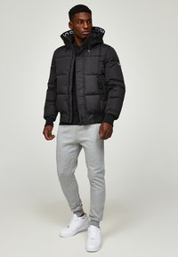 Schwarze Puffjacke mit Kapuze, ausgestattet mit einem steppierten Design und Rippbündchen. Getragen mit hellgrauen Jogginghosen und weißen Turnschuhen.