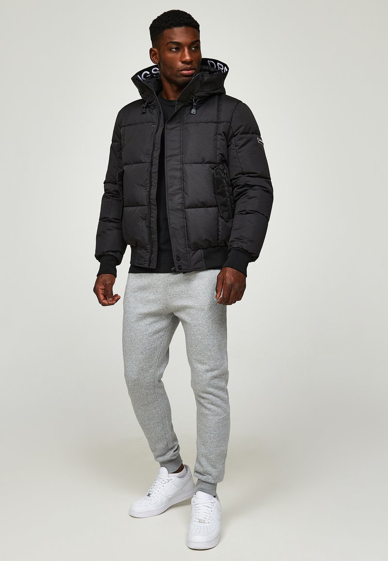 Veste doudoune noire avec capuche, dotée d'un design matelassé et de poignets côtelés. Portée avec un pantalon de survêtement gris clair et des sneakers blanches.