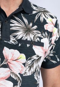 Camisa polo floral en azul oscuro con patrones de hibisco blancos y rosas. Presenta un bolsillo de parche con botones, mangas cortas y cuello con solapa.