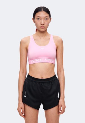 Γυναίκα που φοράει ροζ αθλητικό σουτιέν Nike και μαύρα αθλητικά σορτς Nike, στέκεται με χαλαρά χέρια μπροστά σε απλό φόντο.