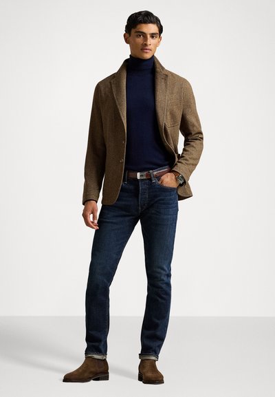 Brauner strukturierter Blazer über einem marineblauen Rollkragenpullover, kombiniert mit dunkelblauen Jeans und braunen Stiefeletten. Inklusive einer Armbanduhr und Gürteldetails.