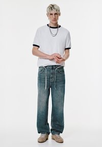Weißes T-Shirt mit kurzen Ärmeln und marineblauem Besatz, locker sitzende, ausgebleichte Jeans und beigefarbene Sneaker. Ein silberner Halskette ist vorhanden.