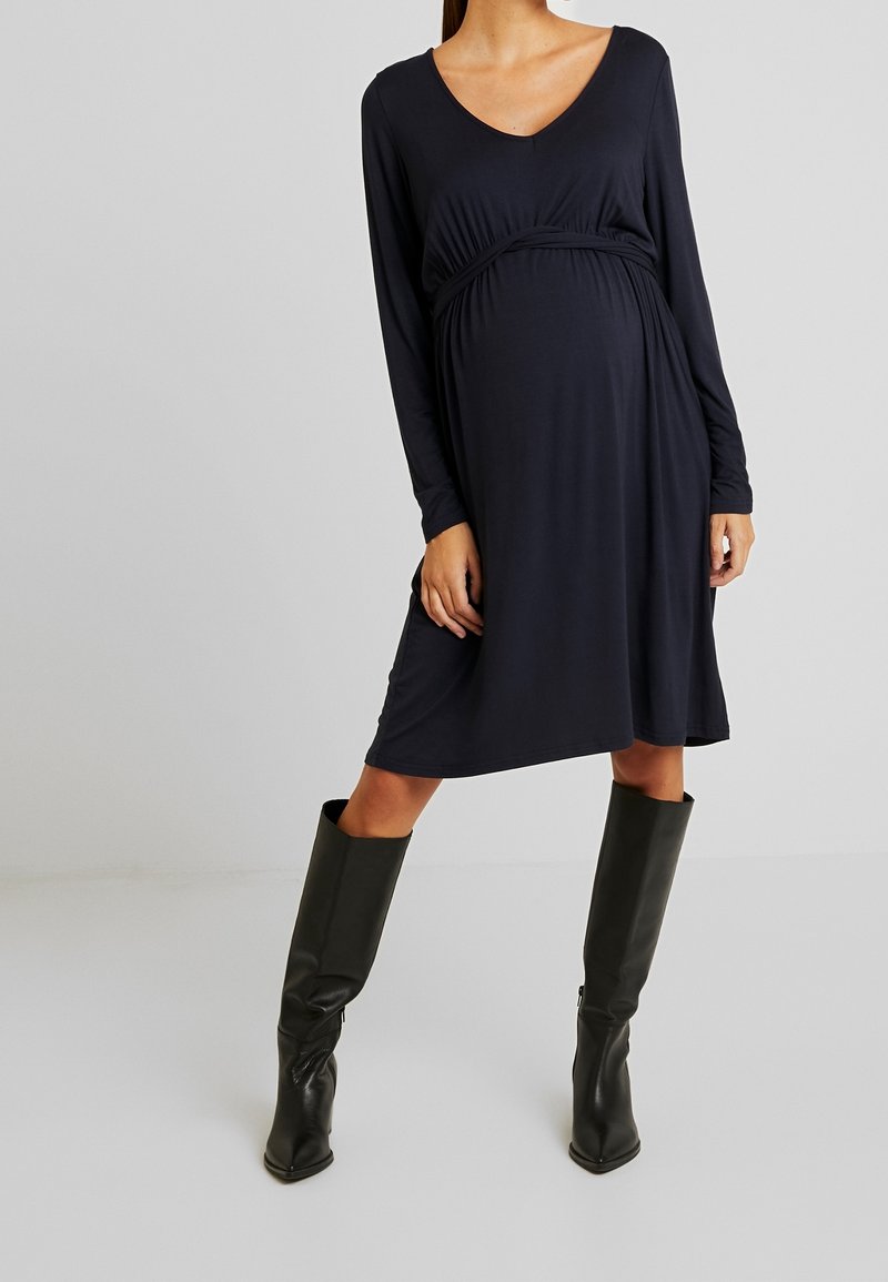 Långärmad, marinblå klänning med V-ringning och rynkad detalj under bysten, kombinerad med knähöga svarta läderstövlar.