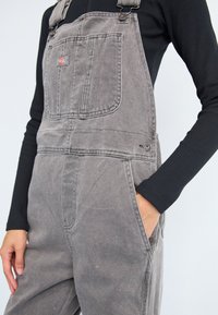 Gray denim overalls with adjustable straps, front pocket detail, and side pockets, featuring a relaxed fit and textured fabric.