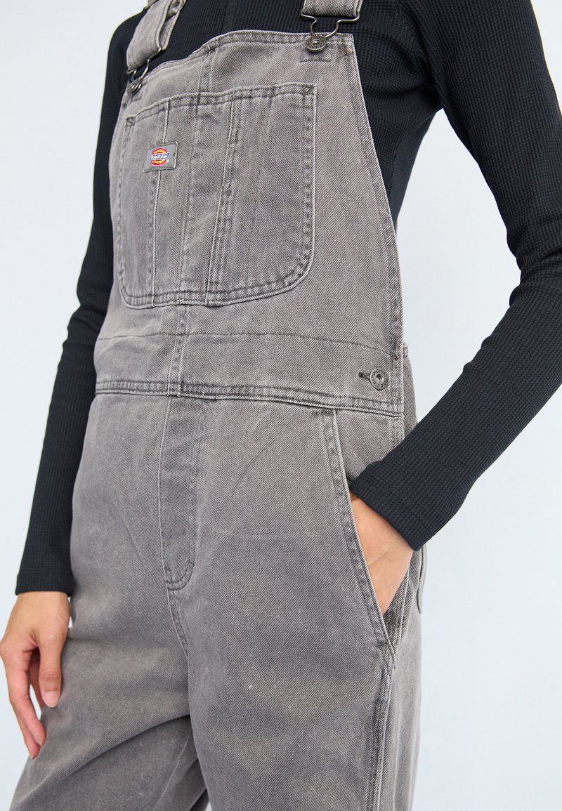 Graue Denim-Latzhose mit verstellbaren Trägern, Fronttasche und seitlichen Taschen, die eine lockere Passform und einen strukturierten Stoff aufweist.