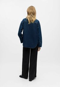 Camicia in denim di colore blu scuro con vestibilità rilassata, maniche lunghe e orlo arrotondato. Abbinata a pantaloni neri a gamba larga e scarpe flat.