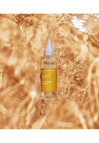 Le toner Essential-C de Murad dans un flacon transparent avec un spray blanc. Le liquide est jaune, entouré d'eau claire ondulante.