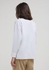 Person mit schulterlangem braunem Haar, der ein langärmliges weißes Shirt und beige Hosen trägt, von hinten gegen einen einfarbigen Hintergrund gesehen.