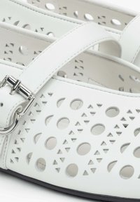 Bílé boty vyrobené z hladké kůže s vyřezávanými kruhovými a trojúhelníkovými vzory, s páskem na stříbrnou sponu a černou podrážkou.