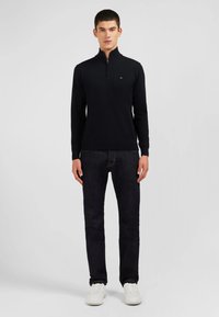 Sweat à zip noir avec manches longues, fabriqué en matériau lisse, porté sur un jean foncé et des baskets blanches. Design simple, logo visible sur la poitrine.