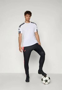 Witte sport T-shirt met zwarte strepen op de schouders, gecombineerd met zwarte broeken met rode accenten. Het model staat naast een voetbal.