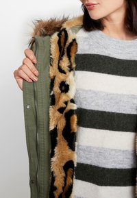 Olive green parka with a leopard print faux fur lining, complemented by a striped gray and green sweater. Close-up of zipper and fur detail.