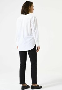 Chemise blanche à boutons avec un ourlet arrondi et des manches longues, associée à un jean slim noir et des mocassins noirs. Texture de tissu lisse.