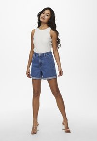 Top de tirantes blanco ligero con textura acanalada, combinado con pantalones cortos de mezclilla azules con dobladillos enrollados. El modelo lleva sandalias de tacón transparentes.