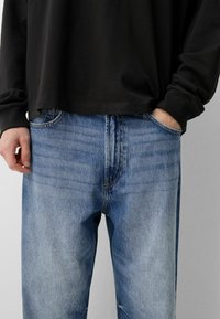 Modri kavbojki s sproščenim fitom, ki imajo rahlo zbledelost in svetle šive. V kombinaciji z črno majico z dolgimi rokavi.