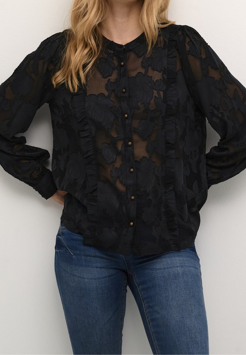 Femme portant un chemisier noir transparent brodé de fleurs avec des volants et des boutons dorés, associé à un jean en denim bleu.