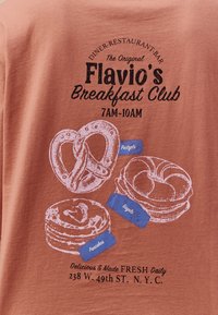 Camisa de color naranja óxido con ilustraciones gráficas blancas de pretzels, bagels y panqueques con etiquetas azules y texto negro para un diner.