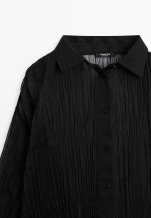 Chemise noire à col pointed, en tissu léger texturé, avec des boutons noirs et un aspect froissé.