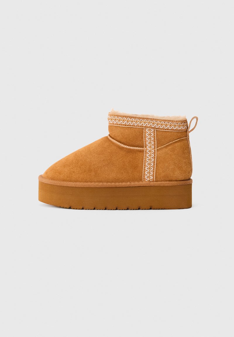 Stivale alla caviglia in suede tan con suola spessa in gomma, cuciture decorative, linguetta a anello e fodera soffice visibile sul bordo superiore.