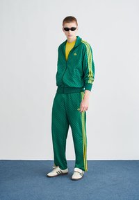 Tuta verde con motivo geometrico, accenti gialli e design a tre strisce. Scarpe da ginnastica bianche completano il look sportivo. Occhiali da sole neri indossati.