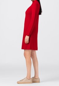 Robe tricotée rouge à manches longues et col montant, avec une coupe droite qui tombe au-dessus du genou, associée à des baskets beiges.