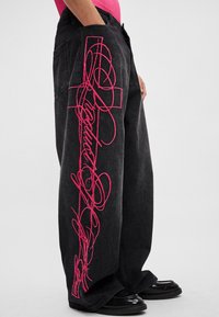 Weite schwarze Denim-Jeans mit leuchtend pinker Stickerei an der Seite, die eine Mischung aus fließenden Mustern und Text zeigt. Strukturierter Stoff mit lockerem Sitz.