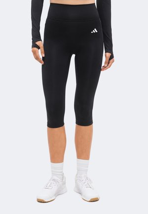 Osoba nosząca czarne legginsy do połowy łydki z logo Adidas, czarną bluzkę na długi rękaw z otworami na kciuki, białe skarpetki oraz białe buty sportowe, stojąca.