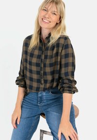 Groen en marineblauwe geblokte blouse met een knopenfront en opgerolde mouwen, gecombineerd met blauwe jeans, zittend op een houten kruk.
