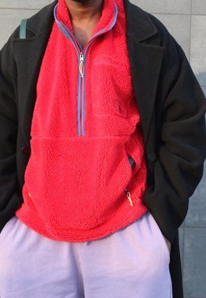 Personne portant un pull en polaire rouge vif avec un manteau noir et un pantalon violet clair, mains dans les poches, debout contre un mur carrelé.
