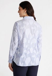Camicia azzurra chiara con motivo floreale bianco, maniche lunghe, colletto con bottoni e spacco posteriore. Realizzata in tessuto leggero con una finitura testurizzata.