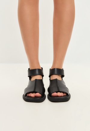 Jambes portant des sandales noires à bouts ouverts avec des brides épaisses à la cheville, debout sur une surface claire avec un fond uni.