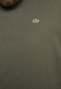 Tessuto verde oliva con un piccolo logo di coccodrillo ricamato in verde, con la bocca aperta e un contorno bianco, nella parte superiore centrale.
