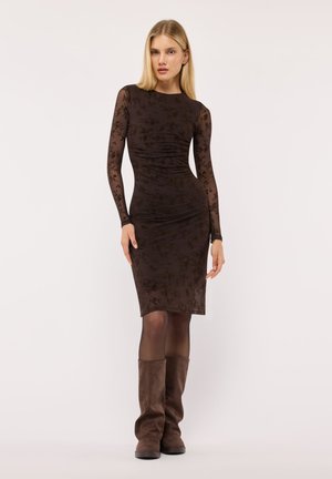 Donna bionda che indossa un vestito nero aderente a fiori trasparente a maniche lunghe, collant scuri e stivali marroni in camoscio al ginocchio.