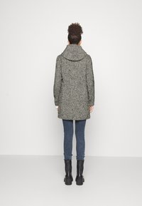 Cappotto grigio e nero a fantasia con un ampio colletto, design di lunghezza media, abbinato a jeans blu e stivali neri massicci. La texture è tessuta.
