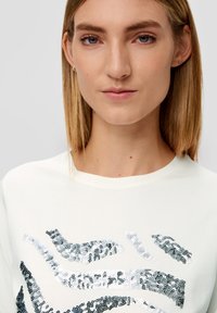 T-shirt couleur crème avec un design en sequins argentés et noirs, col rond et tissu doux avec une texture lisse.