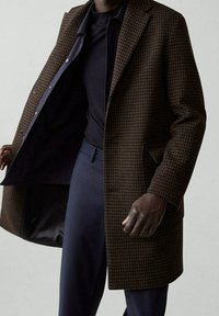 Hnědý kabát s kostkovaným vzorem houndstooth, se střihem na míru, klopami a knoflíkovým zapínáním, oblečený přes námořnické tričko a kalhoty.